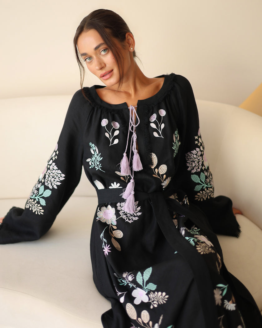 Black linen kaftan maxi dress with pastel floral embroidery and lilac tassel ties styled seated on white sofa - Zalxndra Resort