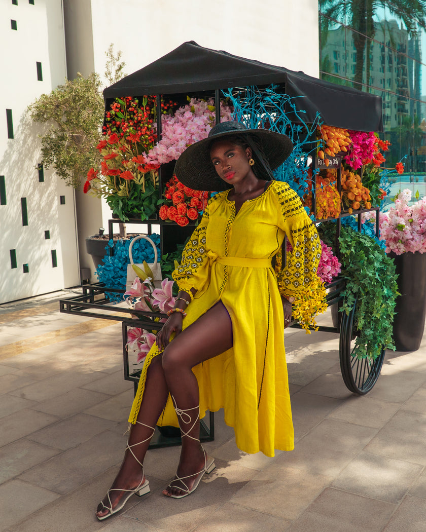 Lemon yellow linen kaftan maxi dress with black floral embroidery, balloon sleeves and self-tie belt, styled with wide-brim hat, editorial seated view - Zalxndra Resort