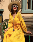 Saffron yellow linen kaftan maxi dress with navy geometric embroidery, balloon sleeves and tassel tie, styled with straw hat and sunglasses, editorial seated view - Zalxndra Resort
