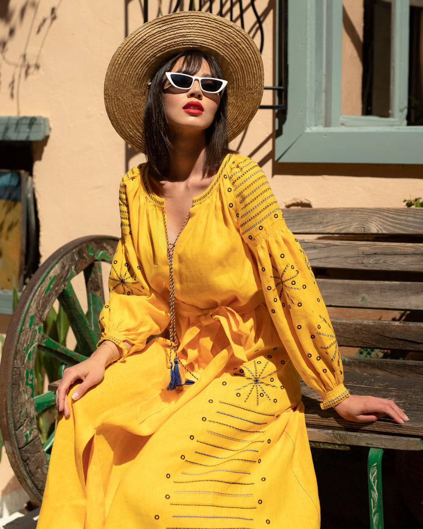 Saffron yellow linen kaftan maxi dress with navy geometric embroidery, balloon sleeves and tassel tie, styled with straw hat and sunglasses, editorial seated view - Zalxndra Resort