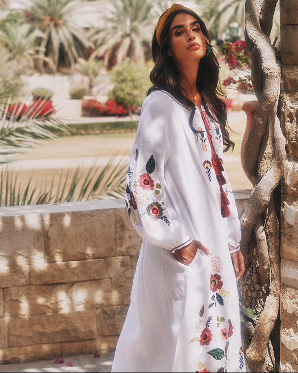 White linen kaftan maxi dress with multicolour folk floral embroidery, balloon sleeves, tassel ties, and side pockets, editorial garden setting - Zalxndra Resort